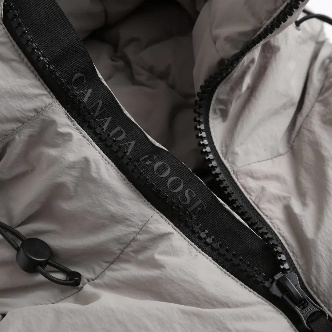 P980 Canada Goose hooded down jacket Fabric selection: Weaved with 400T high-count yarn. After special water-repellent treatment, the surface forms a lotus leaf effect - raindrops will slide down in beads, and snow particles will fall off when touched. The tear resistance reaches 200N (approximately 20kg load-bearing), the light gray matte texture breaks the stereotype of "outdoor clothing = hardcore", and the soft luster presents a high-end matte effect under urban neon lights, seamlessly switching scenes from ski resorts to CBD office buildings.  Process 1: Use polyester woven ribbons as the base, and use high-precision computer embroidery to decorate the brand letters on the ribbons. The edges of the pattern are clear, the stitches are fine and even, and multiple layers of embroidery threads are stacked to enhance the three-dimensional effect.  Process 2: Precision thread reinforcement to prevent velvet drilling and ensure that the fluffy and resilient elasticity lasts as new.  Details: The embedded cuffs are three-dimensionally woven to form a "wavy" elastic structure, forming natural folds when fitting the wrist, increasing the contact area with the skin, thus improving the windproof effect. At the same time, micro-velvet treatment is added to the inside of the cuffs, which feels like a second layer of skin and avoids friction on the skin from metal zippers or hard fabrics.  90 national standard high-quality white duck down: selected high-loft down, weighing 240g, light and warm, with excellent temperature locking performance and good resilience.  Accessories: 1:1 customized armband, 1:1 customized zipper pendant, 1:1 customized elastic buckle, etc. SIZE: S M L XL